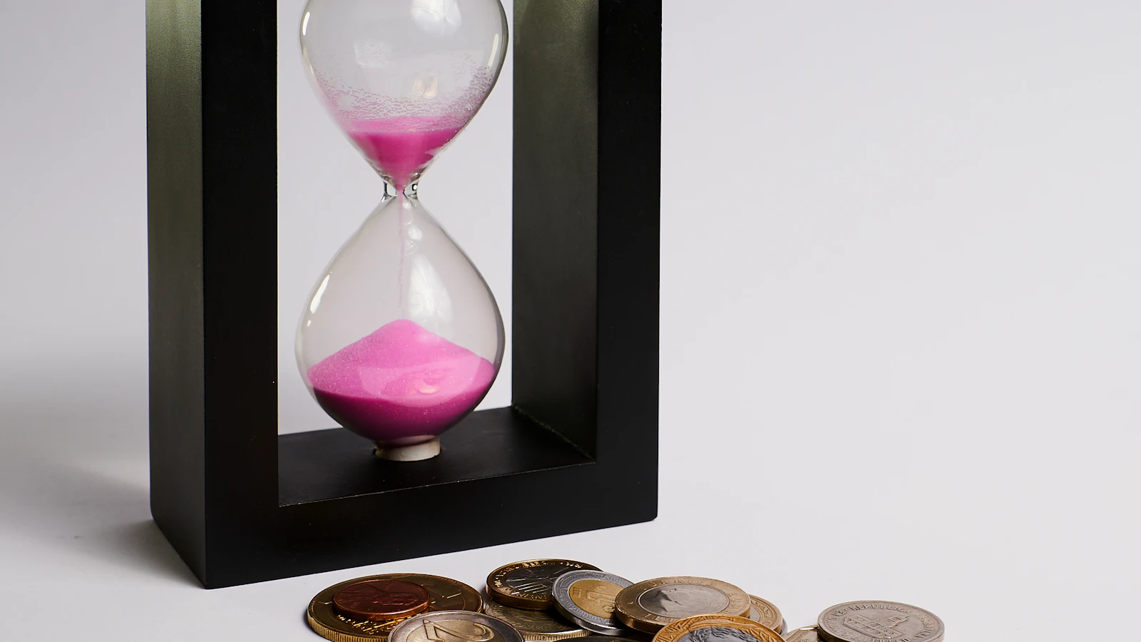 Een zandloper met roze zand naast een stapel euromunten, symbool voor sparen doorheen de tijd