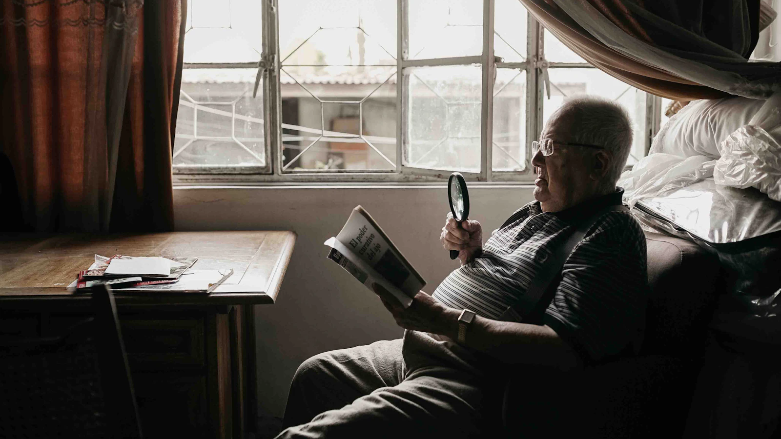 Een oudere man leest thuis in een fauteuil een document met een vergrootglas